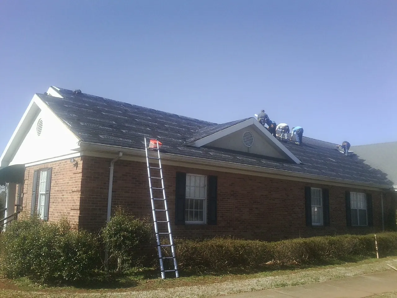 Crew tearing shingles off a brick ranch roof with a ladder set — Emergency Roof Repair work by our crew in Colgate