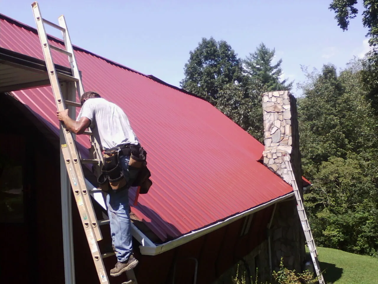 Roofer on a ladder beside a red metal roof and stone chimney — Soffit & Fascia Repair by Roofing Brothers in Colgate